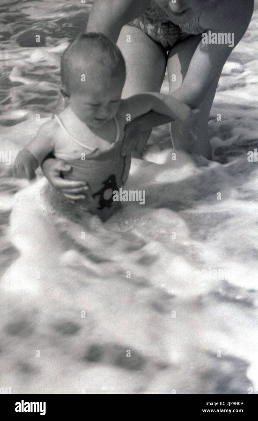 1950, historique, dans l'eau de shallaow, étant tenu doucement par son mousseur, un enfant en bas âge obtenant sa première expérience de la mer, Angleterre, Royaume-Uni. Banque D'Images