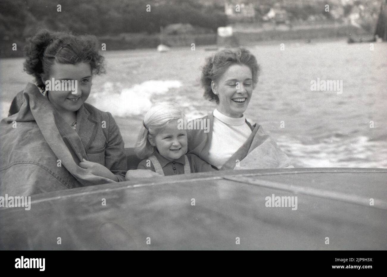 1950, historique, mère avec des filles assis dans un hors-bord., imperméable couvrant dessus, arrêtant le jet de la mer, Angleterre, Royaume-Uni. Banque D'Images