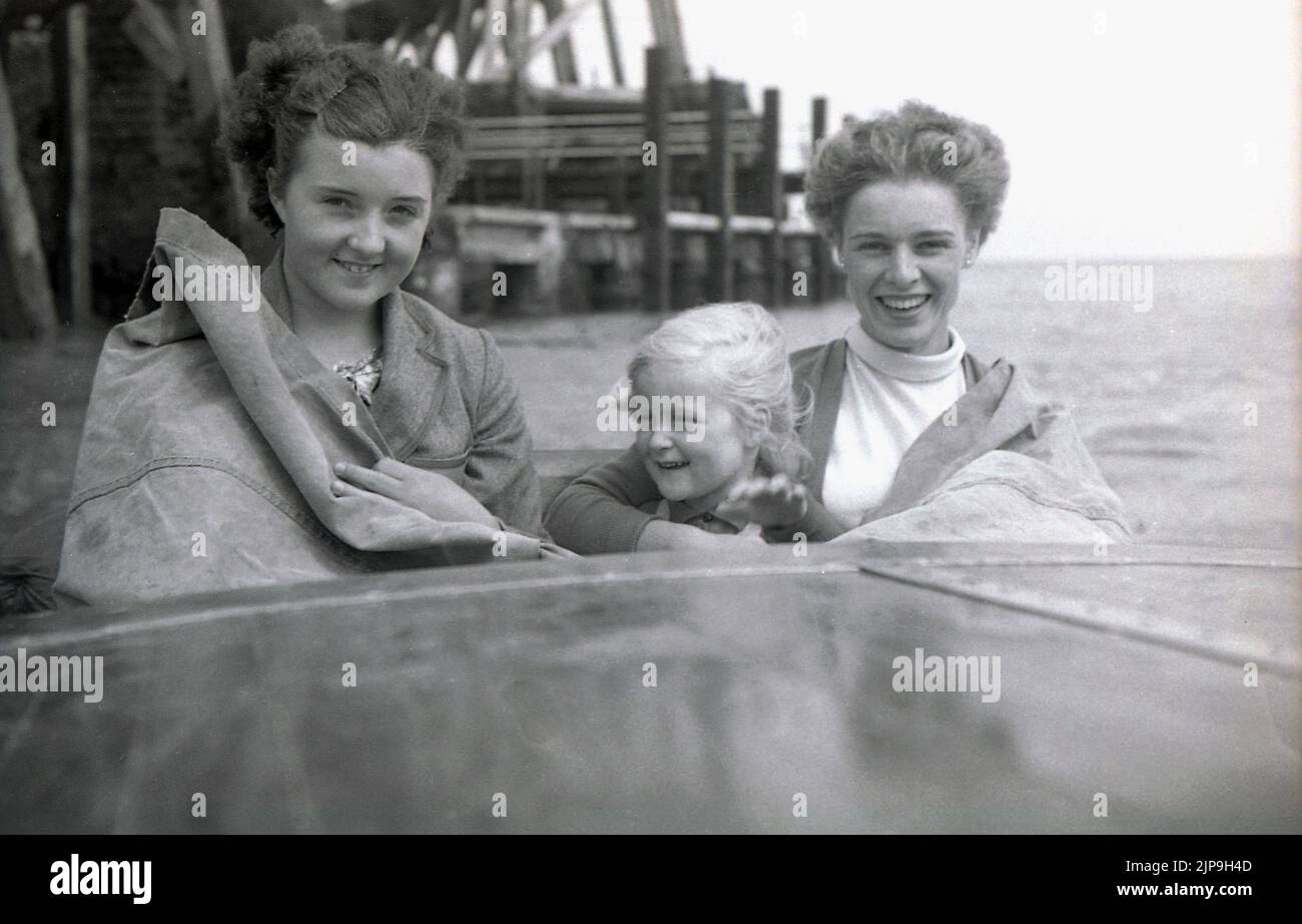 1950, historique, mère avec des filles assis dans un hors-bord., imperméable couvrant dessus, arrêtant le jet de la mer, Angleterre, Royaume-Uni. Banque D'Images
