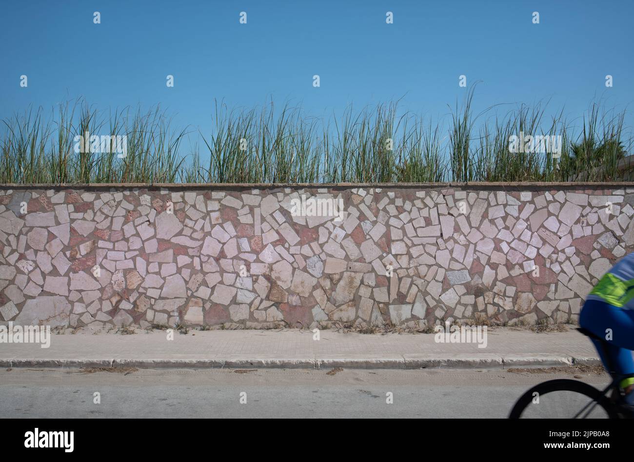 L'arrière d'un cycliste peut être vu au bord de l'image. Au-delà est un mur de pierre, une route, l'herbe poussant sur le mur contre un bleu Banque D'Images
