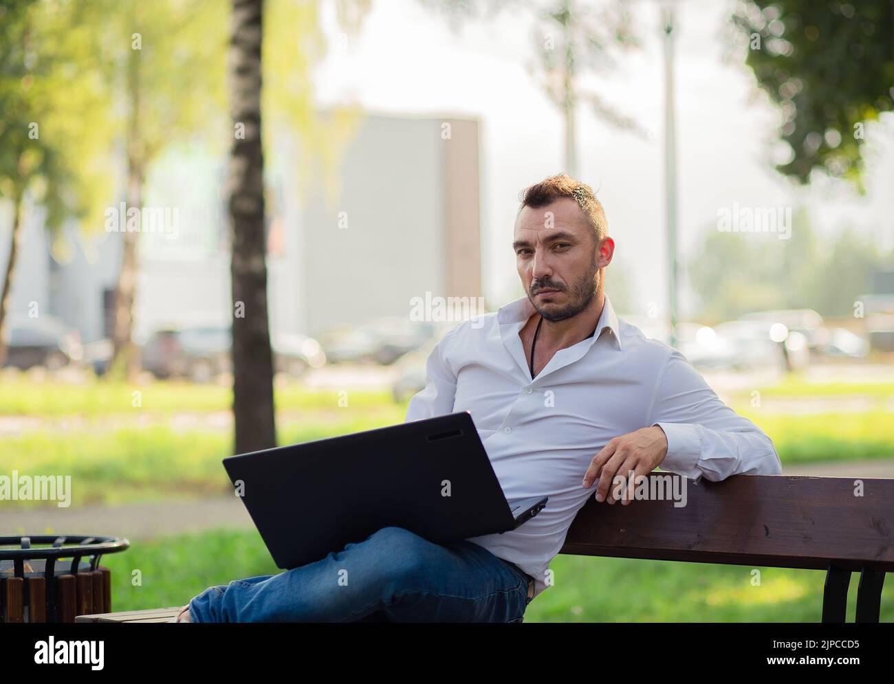 Un beau homme d'affaires dans une chemise blanche travaille dans le parc avec un ordinateur portable. Un jeune homme sur fond d'arbres verts, une chaude journée d'été ensoleillée. Lumière douce et chaude, gros plan. Banque D'Images