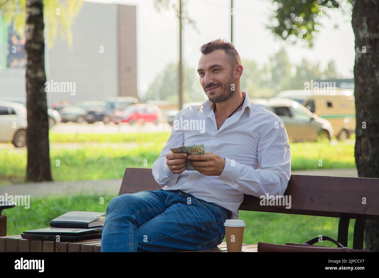 Un homme heureux est assis sur un banc dans le parc, compte tenu des bénéfices. Un jeune homme sur fond d'arbres verts, une chaude journée d'été ensoleillée. Lumière douce et chaude, gros plan. Banque D'Images