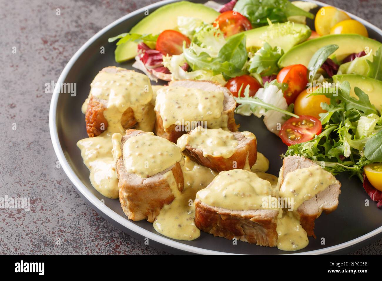Des morceaux de filet de porc tendre servis avec de la sauce à la moutarde au miel et de la laitue, de l'avocat et de la salade de tomates dans une assiette sur la table. Horizontale Banque D'Images