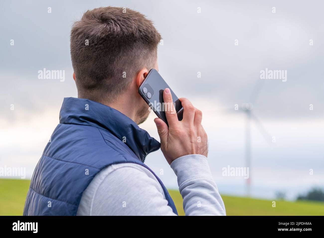 Un ingénieur signale le fonctionnement de l'éolienne via un smartphone en regardant le moulin à vent. L'éolienne produit de l'énergie verte contre le ciel gris Banque D'Images