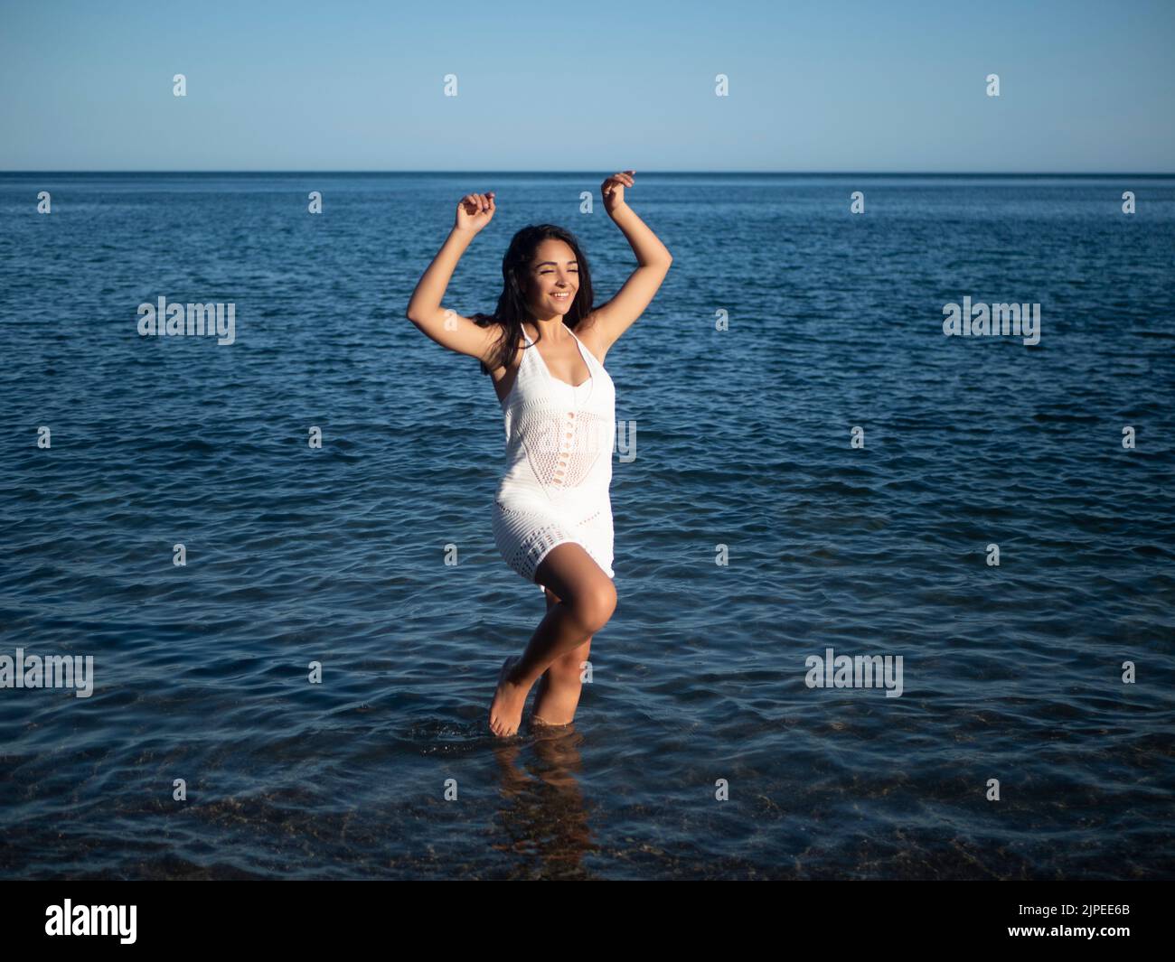 Pleine longueur de contenu jeune hispanique femme voyageur avec de longs cheveux foncés en crochet blanc robe marchant dans l'eau peu profonde de mer et souriant pendant le summe Banque D'Images