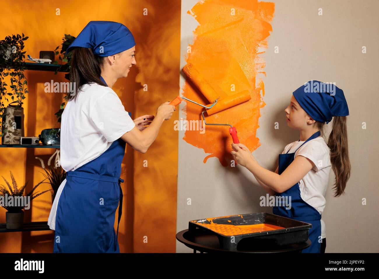 Un petit enfant aide maman à peindre les murs, en utilisant de la peinture orange pour rénover et redécorer la chambre de la maison. Appartement jeune famille rénove avec rouleau de pinceau et outils, travaux ménagers. Banque D'Images
