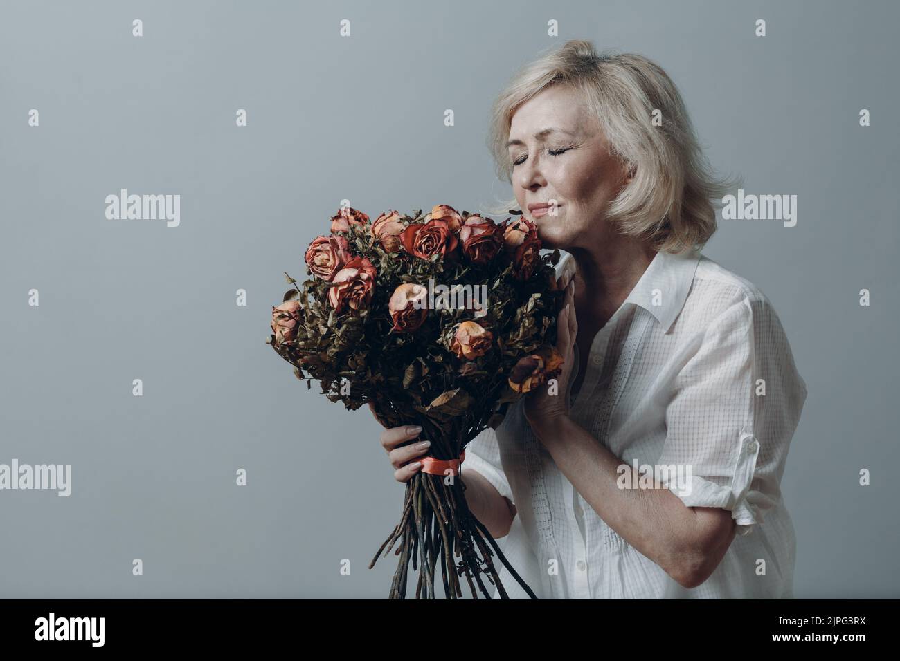 Triste femme âgée touchant un bouquet de fleurs de rose flétrisées. Femme adulte aux cheveux gris dans la dépression. Concept de vieillissement ancien. Studio Shoot isolé sur fond gris. Copier l'espace Banque D'Images