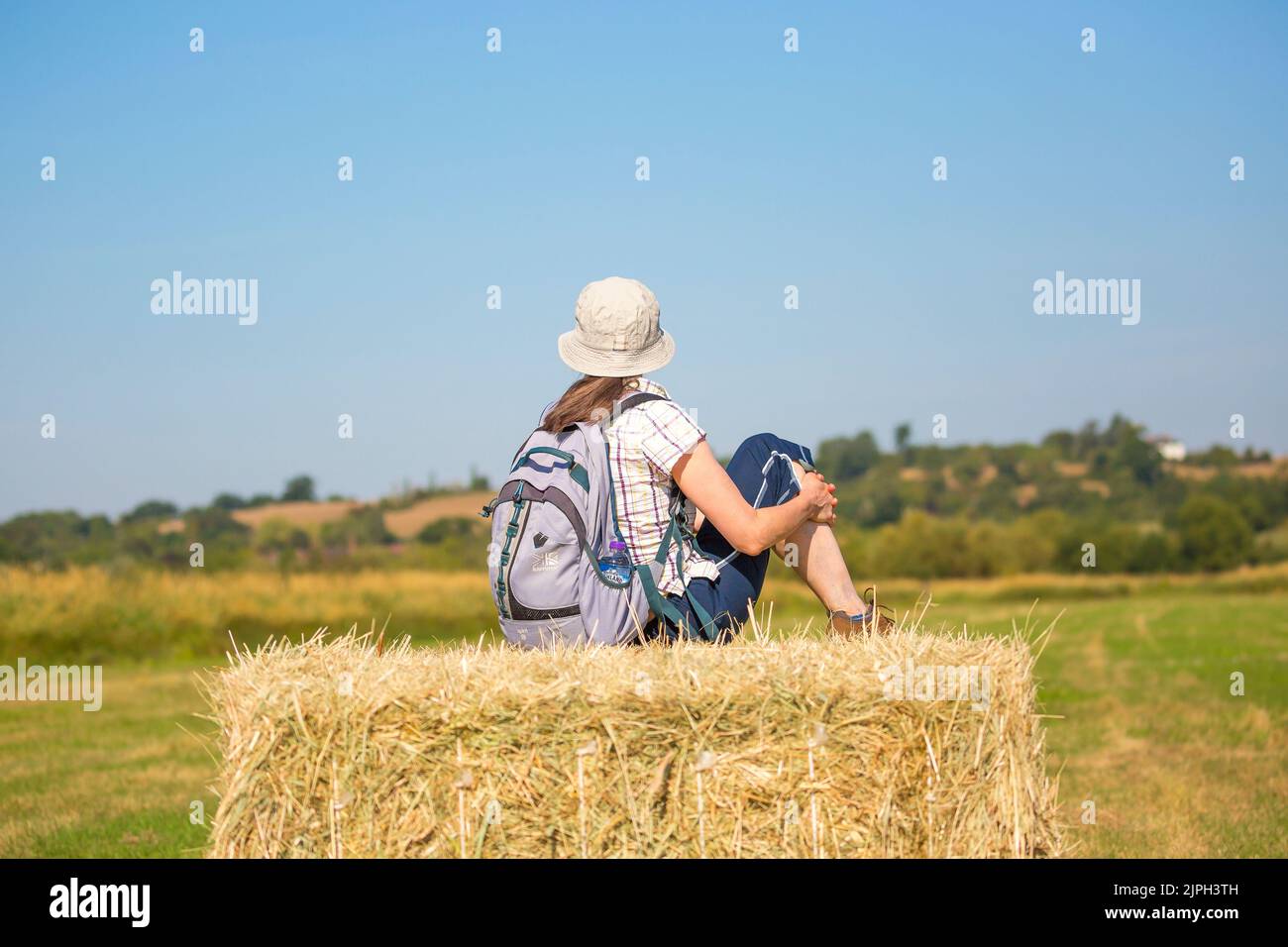 Vue arrière de la rampe femelle isolée sur une balle de paille/foin dans la campagne du Royaume-Uni, avec chapeau de soleil et sac à dos pendant la vague de chaleur estivale du 2022 août. Banque D'Images