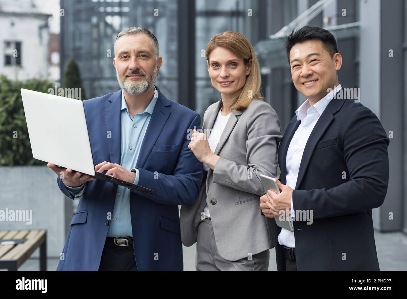 Portrait de trois collègues d'affaires divers, groupe d'affaires à l'extérieur du bâtiment de bureau, homme et femme asiatique regardant l'appareil photo et souriant, équipe de rêve avec ordinateur portable Banque D'Images