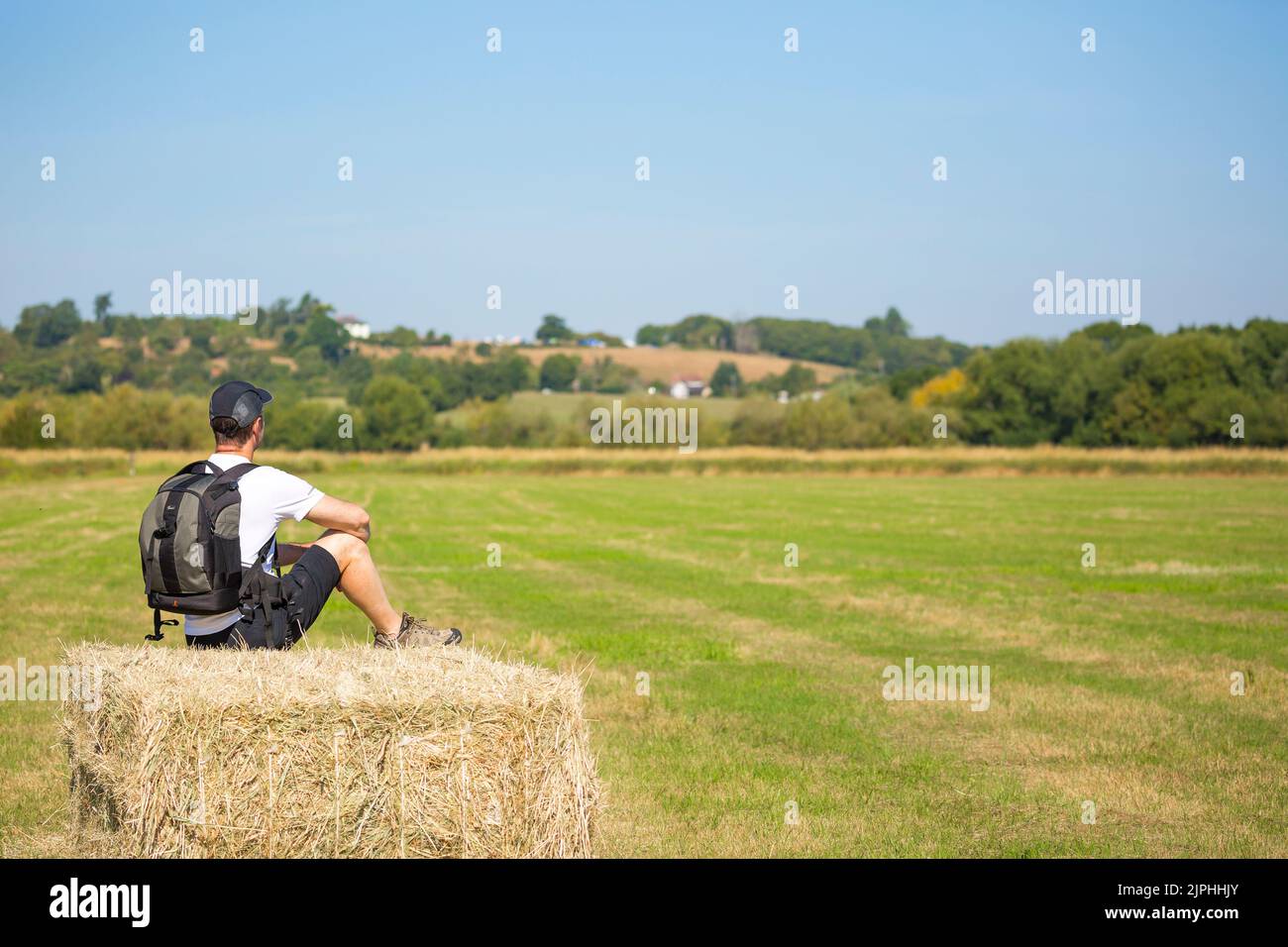 Vue arrière d'un homme en short et casquette, sac à dos à l'arrière, assis isolé sur une balle de paille/foin dans la campagne du Royaume-Uni lors d'une chaude journée d'été. Banque D'Images