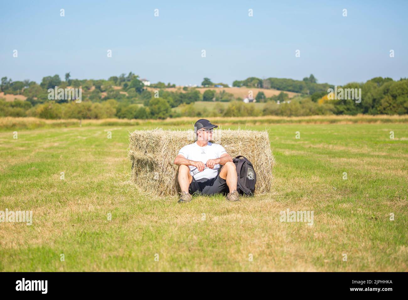 Homme assis isolé par la balle de paille/hale (en short et en chapeau) lors d'une chaude journée d'été, campagne britannique. Banque D'Images