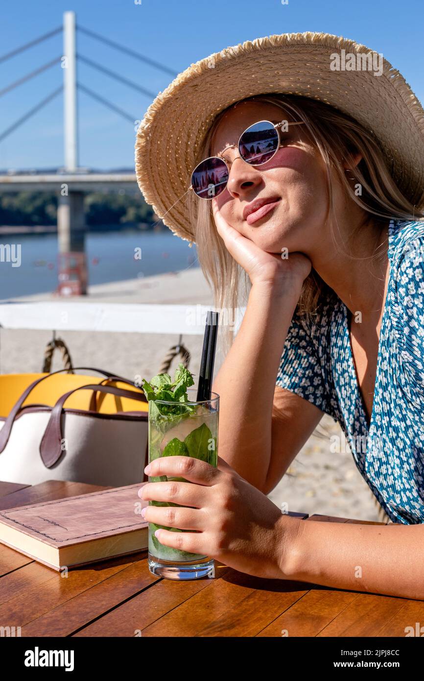 Une belle jeune femme portant un chapeau de paille et des lunettes de soleil, prenant un cocktail et profitant d'un agréable après-midi d'été dans un café de plage au bord de la rivière. Banque D'Images