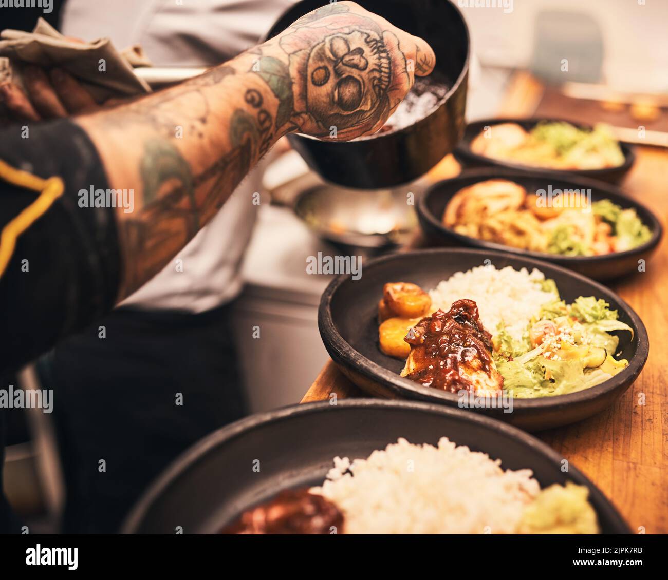 Les touches de finition. Gros plan d'un chef méconnu tatoué les mains servant la nourriture dans des assiettes à l'intérieur d'une cuisine d'un restaurant. Banque D'Images