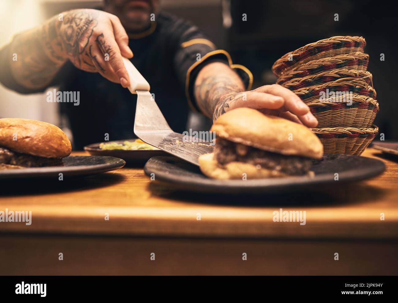 Commandez. Gros plan d'un chef méconnu tatoué les mains servant un hamburger dans une assiette à l'intérieur d'une cuisine d'un restaurant. Banque D'Images