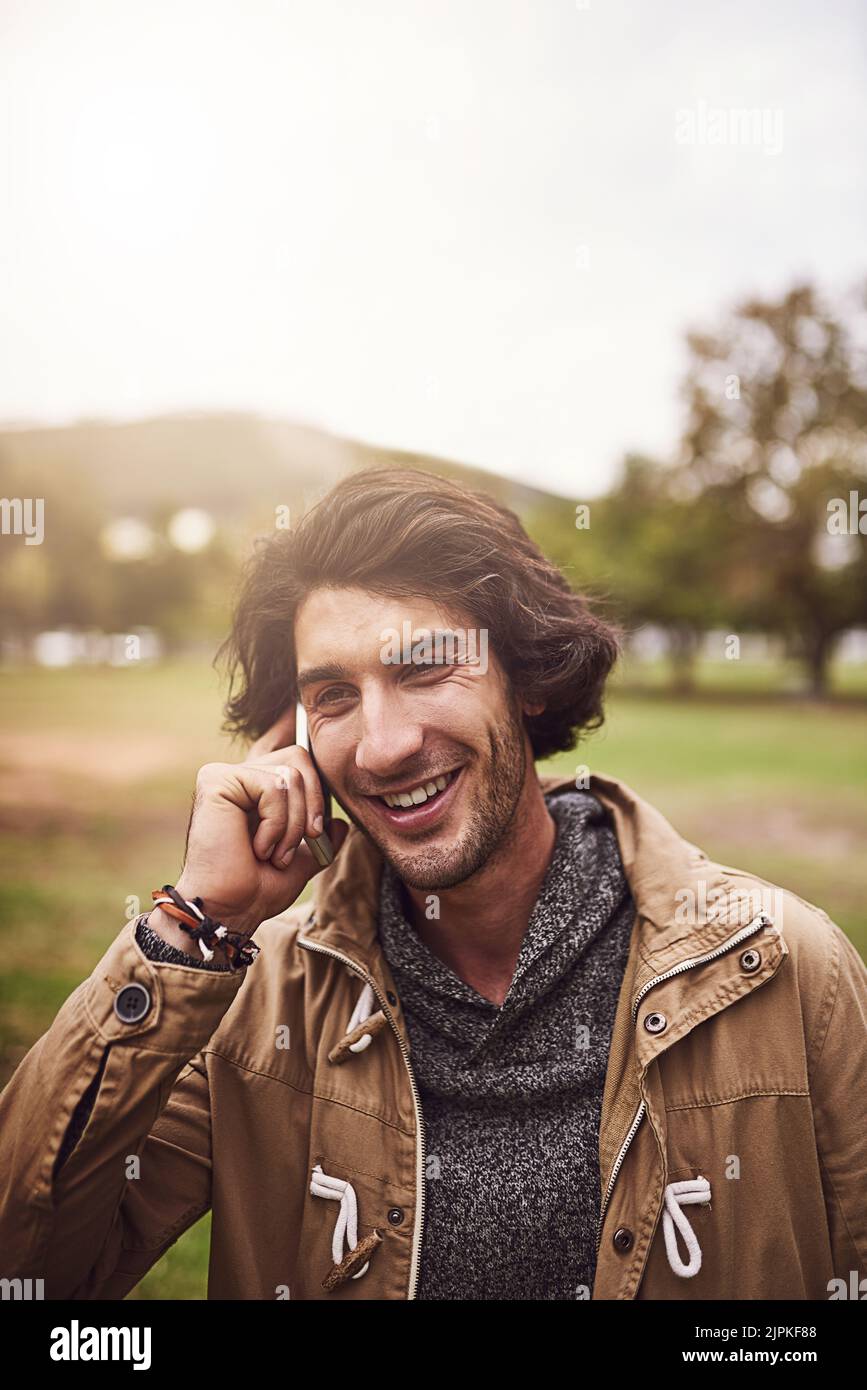 Nous pouvons parler pendant des heures. Un jeune homme joyeux qui parle au téléphone tout en se tenant à l'extérieur dans un parc. Banque D'Images