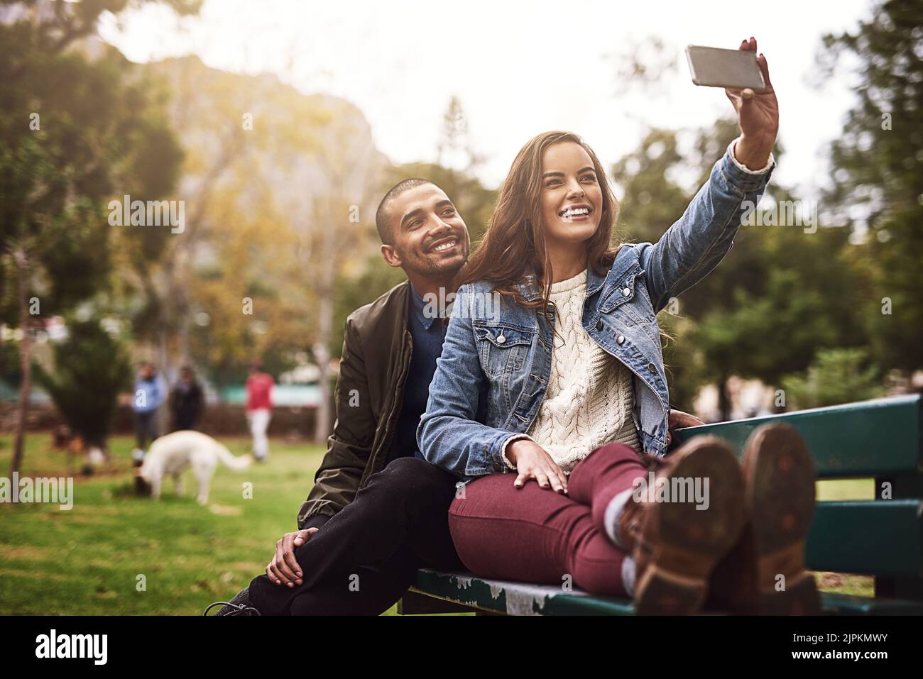 Ne manquez pas d'avoir l'arrière-plan. Un jeune couple gai assis sur un banc tout en prenant des autoportraits ensemble à l'extérieur dans un parc. Banque D'Images