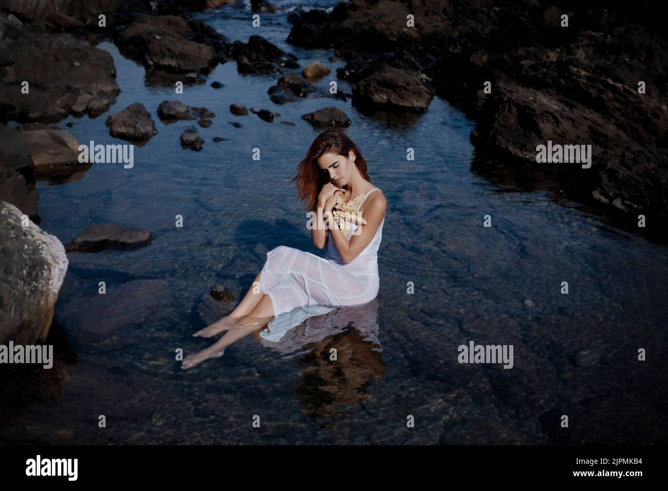Vue latérale d'une jeune femme élégante avec de longs cheveux foncés en robe blanche, assise sur la pierre en mer et embrassant l'étoile de mer dans la nature Banque D'Images