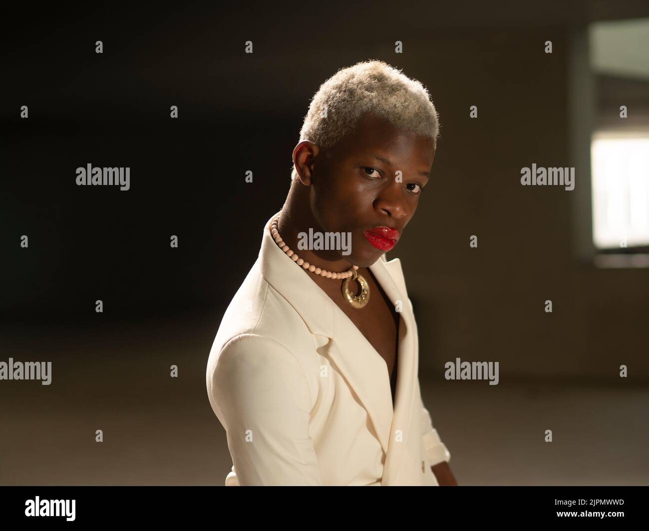 Modèle afro-américain élégant en veste blanche élégante avec cheveux blonds courts et lèvres rouges regardant l'appareil photo avec visage sérieux sur le dos flou Banque D'Images