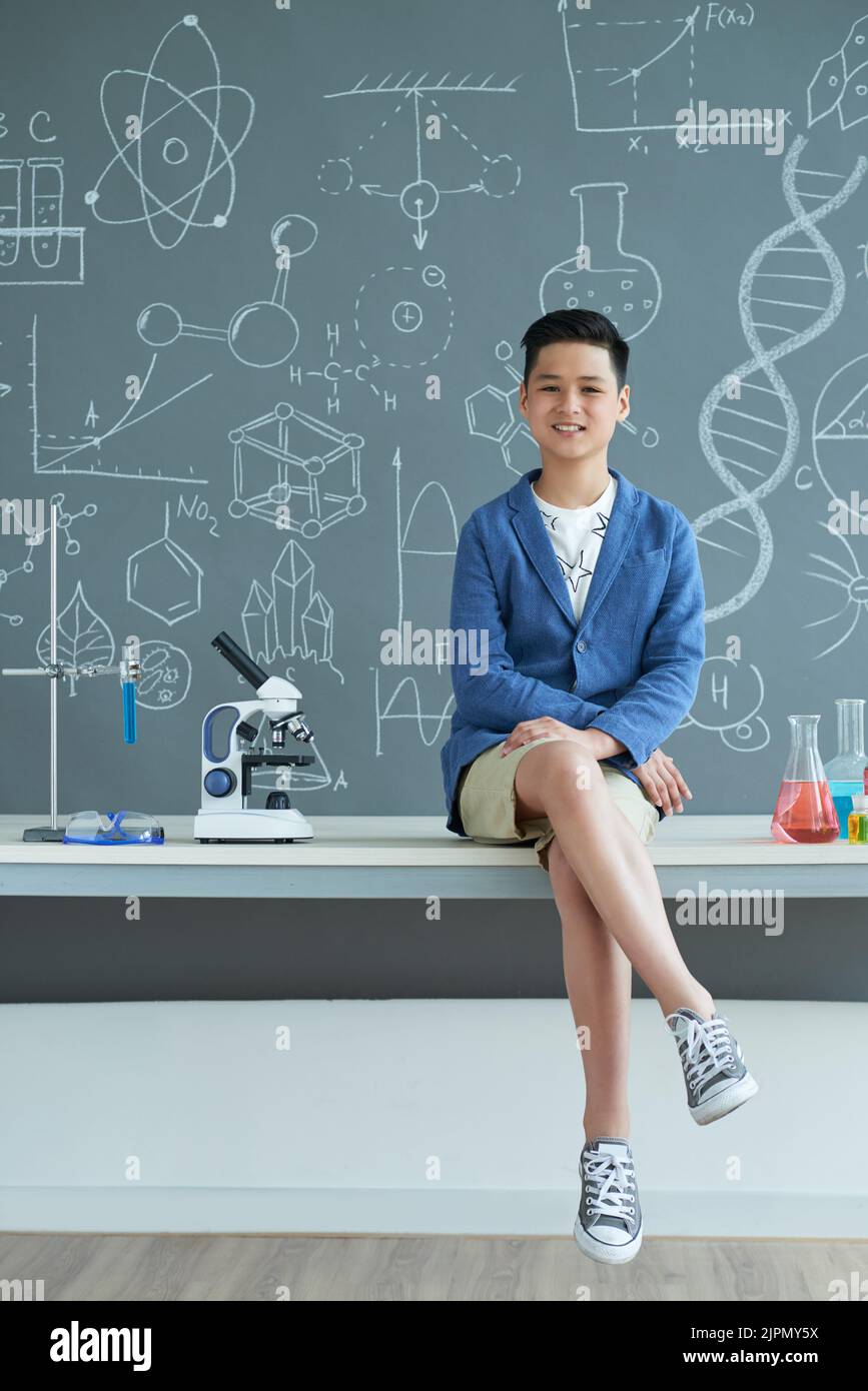 Portrait complet d'un adolescent gai en tenue décontractée assis sur un bureau avec les jambes croisées et posant pour la photographie, à l'intérieur de la salle de classe de chimie moderne en arrière-plan Banque D'Images