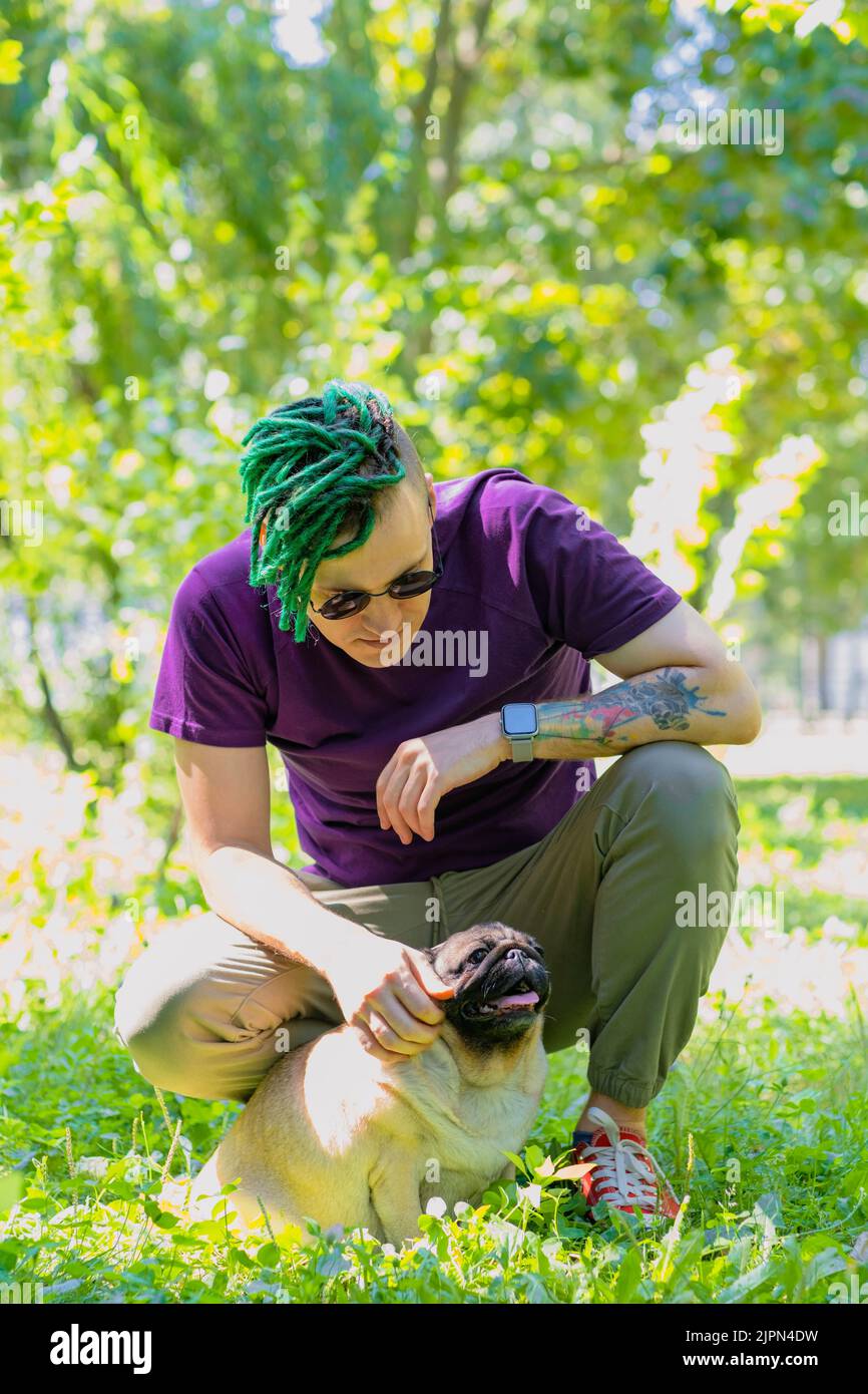 Un hipster avec des dreadlocks verts et des verres ronds noirs est assis sur une pelouse verte et de bourracher un pug mignon lors d'une journée ensoleillée d'été. Un homme marche un pug Banque D'Images