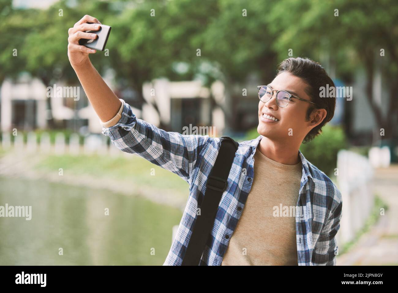 Homme asiatique gai portant des lunettes élégantes et portant un selfie sur un smartphone moderne, tout en profitant d'une agréable promenade dans un parc public, prise de vue portrait Banque D'Images