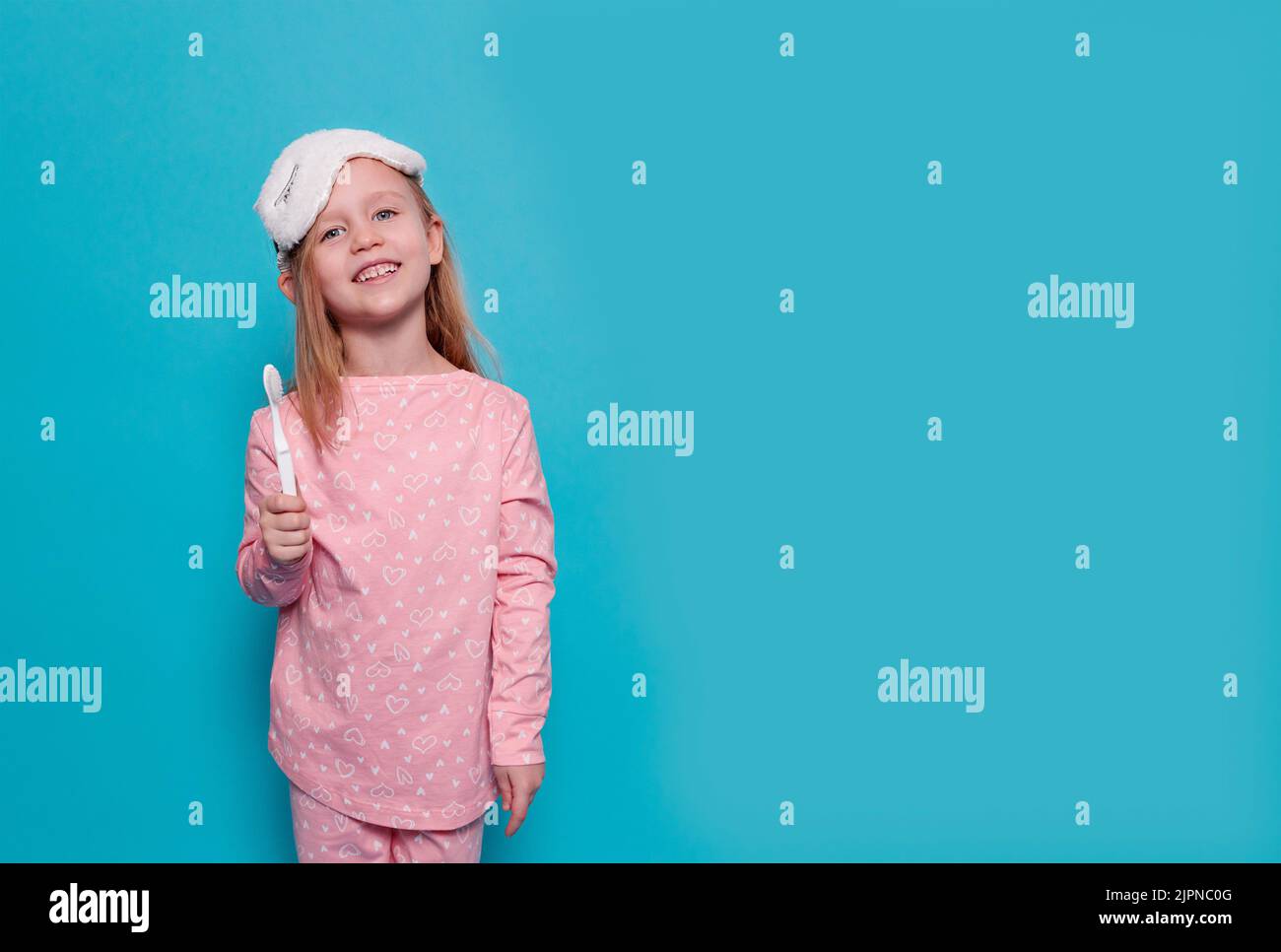 une petite fille en pyjama et un masque de sommeil montre une brosse à dents, sur un fond bleu Banque D'Images