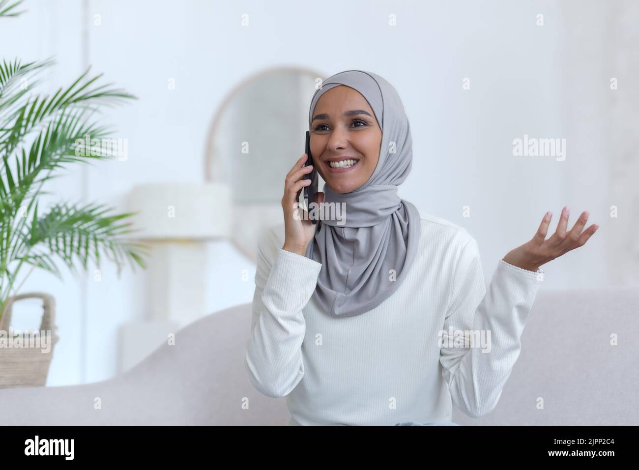 Jeune belle femme musulmane dans le hijab à la maison parlant au téléphone, femme dans le salon souriant et heureux tenant le smartphone à la main bavardant avec des amis Banque D'Images