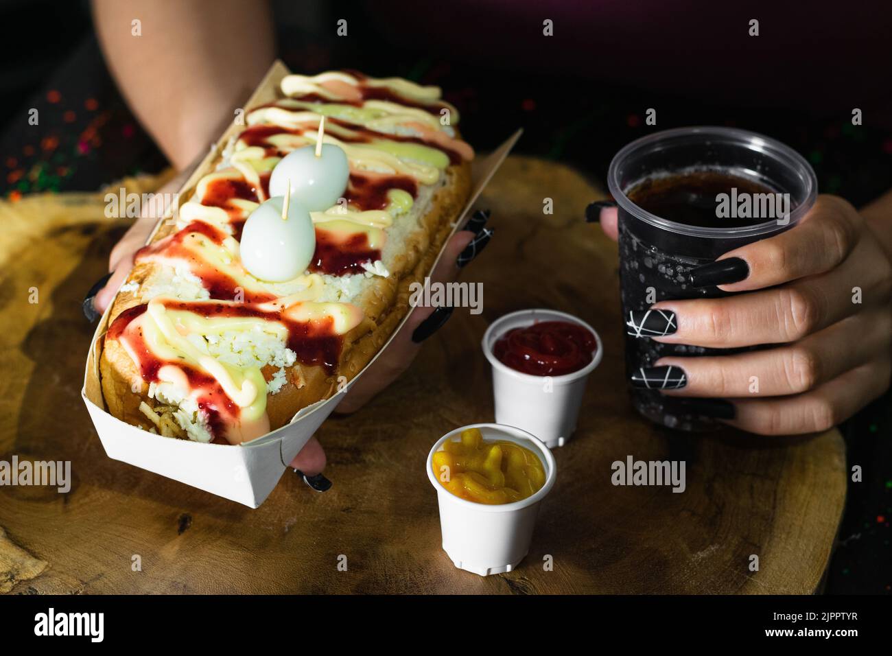 fille tenant dans ses mains un hot dog avec des sauces, du fromage et deux œufs de caille sur le dessus, sombre et fond de bois, à côté de lui il ya deux tasses en plastique w Banque D'Images