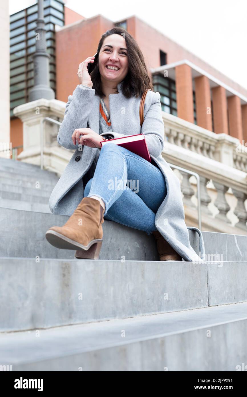 Jeune femme caucasienne souriante ayant une conversation téléphonique. Elle est assise sur quelques marches à l'extérieur. Banque D'Images