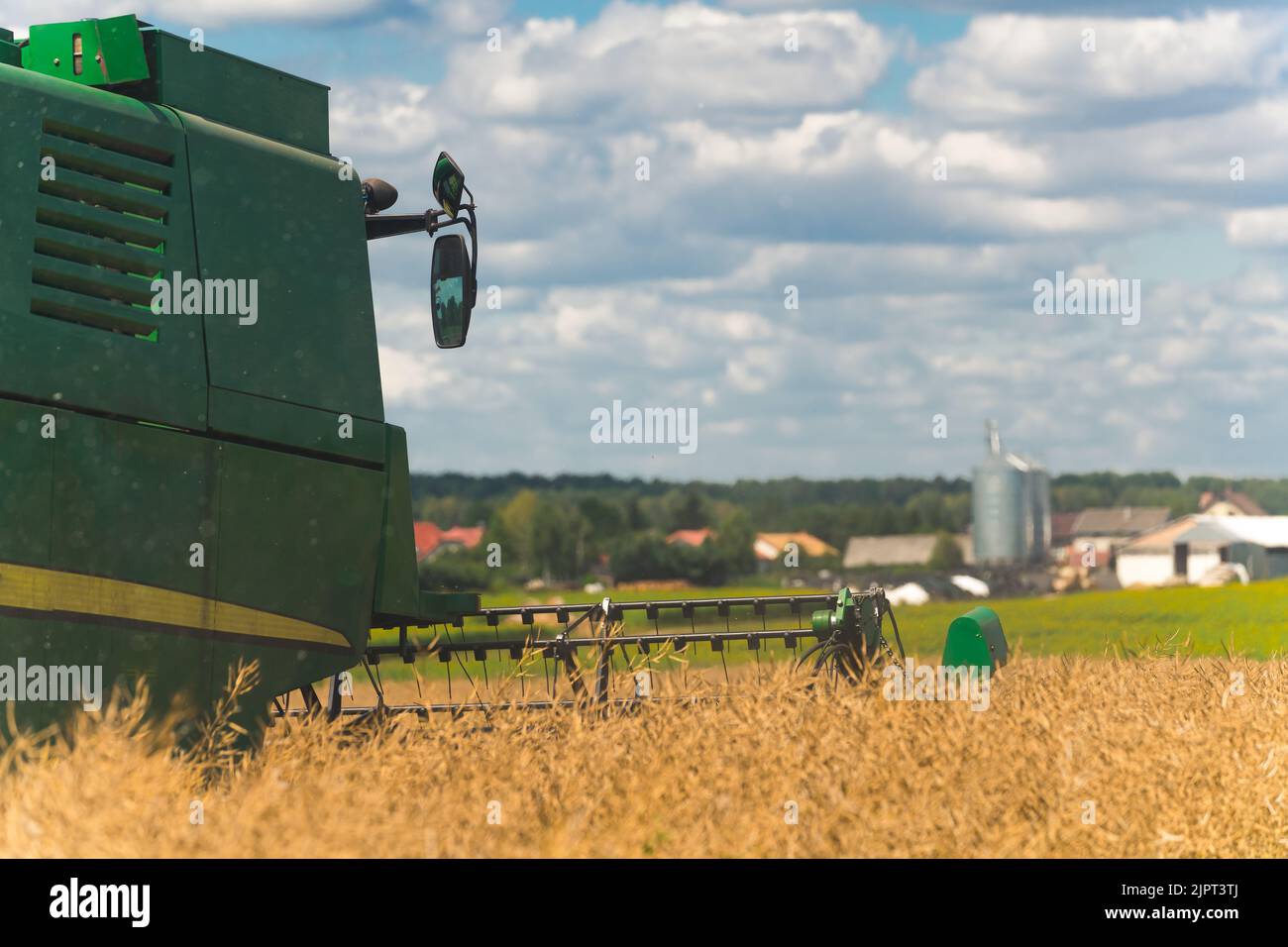 L'arrière de la moissonneuse-batteuse moderne de couleur verte et son rabatteur moissonnant du colza. Travail à col bleu. Maisons de campagne dans le fond flou. Photo de haute qualité Banque D'Images