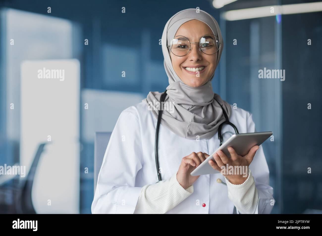 Jeune belle femme asiatique musulman médecin dans hijab souriant et regardant la caméra travailler avec un ordinateur tablette dans le bureau de clinique moderne à l'intérieur du médecin dans une robe médicale et des lunettes. Banque D'Images