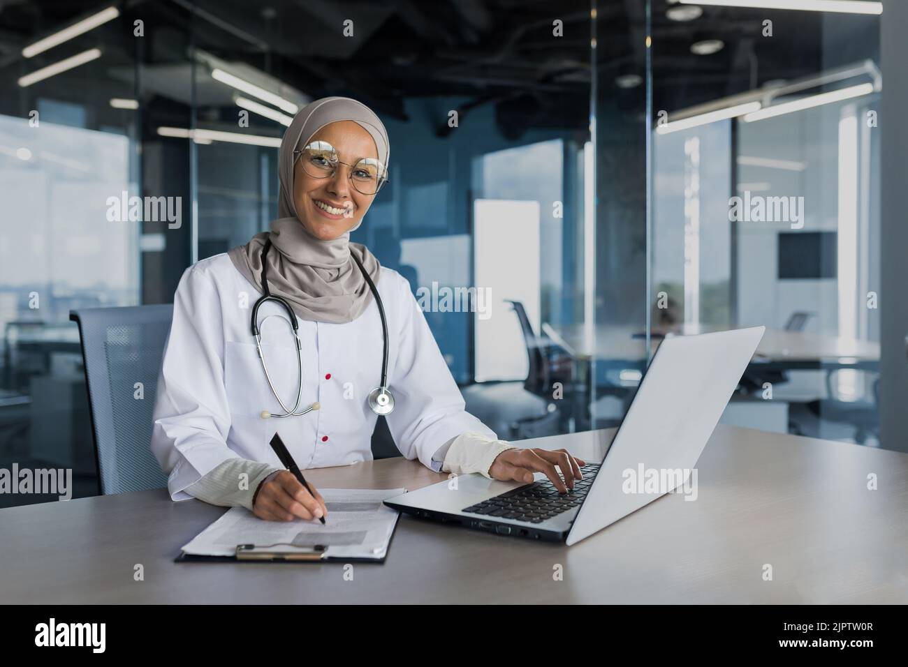 Portrait d'une femme médecin dans le hijab, musulmane travaillant dans un bureau moderne à l'intérieur de la clinique, souriant et regardant l'appareil photo, remplissant des documents, à l'aide d'un ordinateur portable Banque D'Images