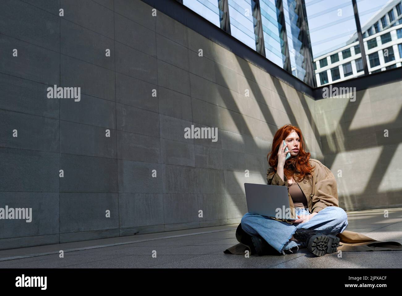 Une jeune fille à tête rouge utilisant un ordinateur portable parlant sur une cellule dans un emplacement urbain. Banque D'Images