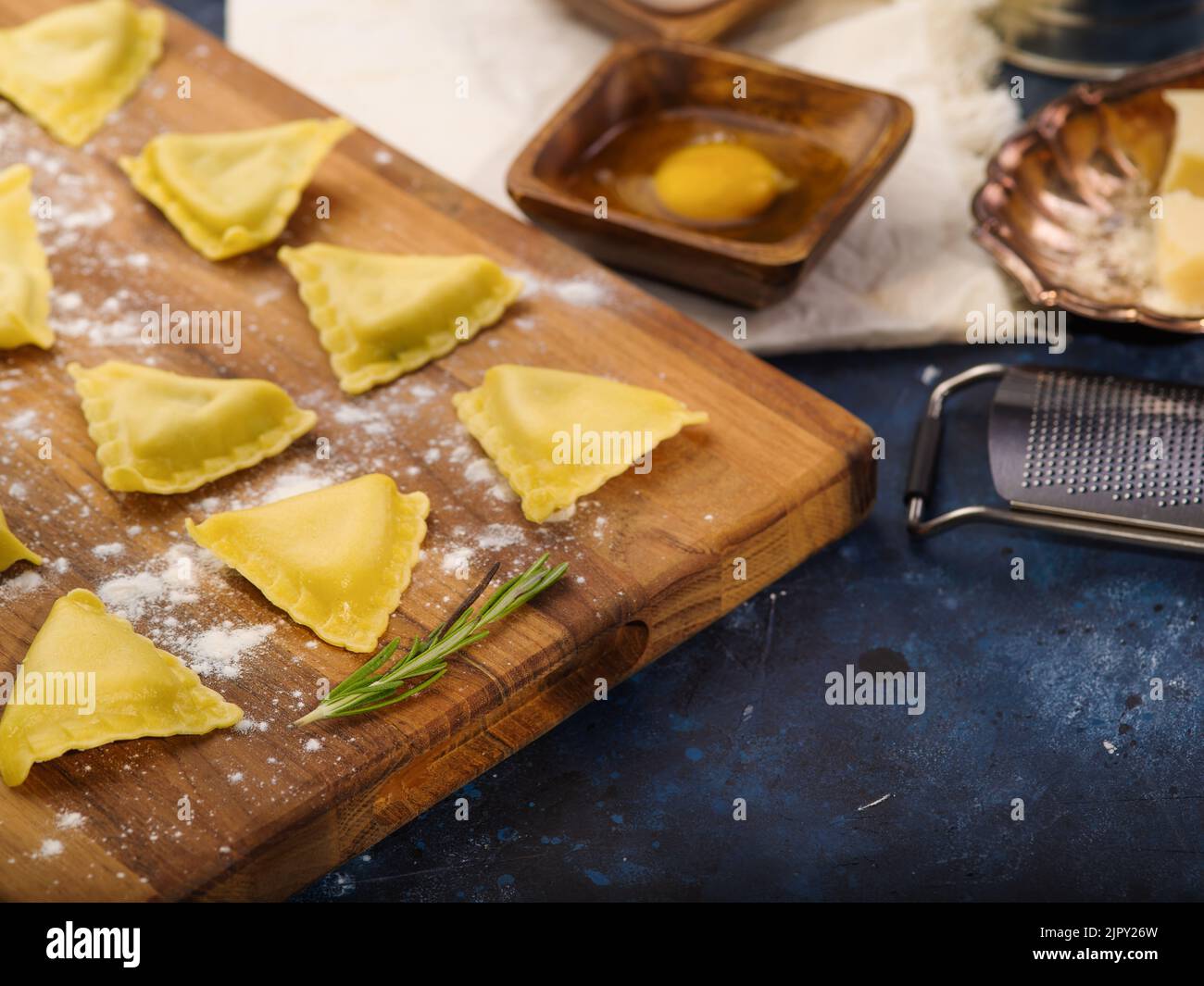 Le processus de fabrication de raviolis faits maison. Raviolis italiens bruts triangulaires sur une planche à découper, ingrédients, ustensiles de cuisine sur fond bleu foncé. Il Banque D'Images