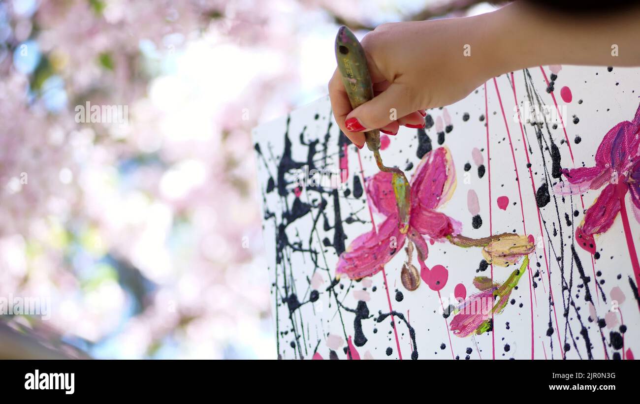 gros plan, femme main, peintre, artiste peint une image de fleurs dans le verger de pomme de printemps en fleur, elle applique des peintures sur toile avec une petite spatule spéciale, tient la palette avec des peintures. Photo de haute qualité Banque D'Images