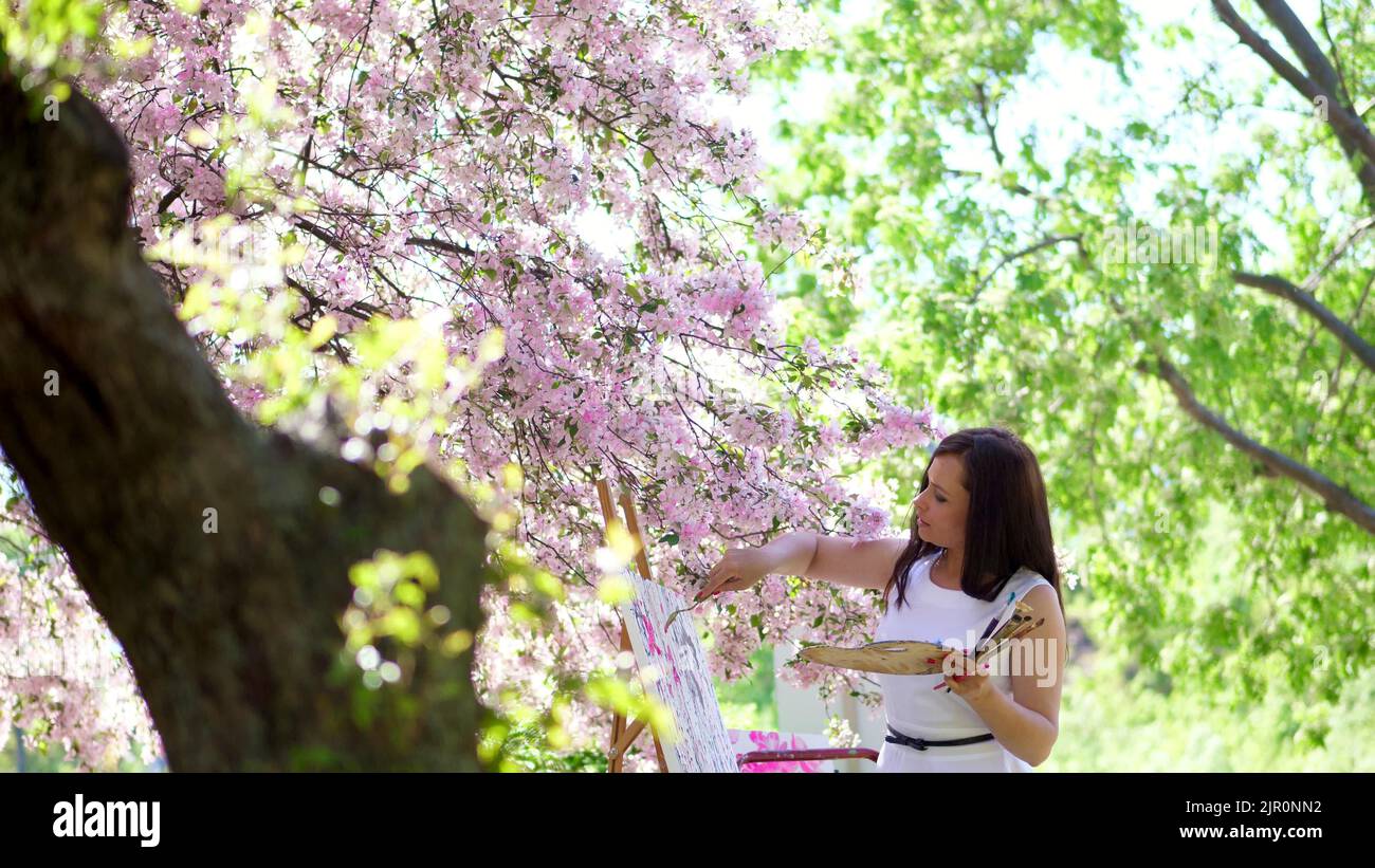 belle femme peintre en robe blanche, artiste peint une image de fleurs dans le verger de pomme de printemps en fleur, elle applique des peintures sur la toile avec une petite spatule spéciale, en utilisant une technique de dessin spéciale. tient une palette avec des peintures. Photo de haute qualité Banque D'Images
