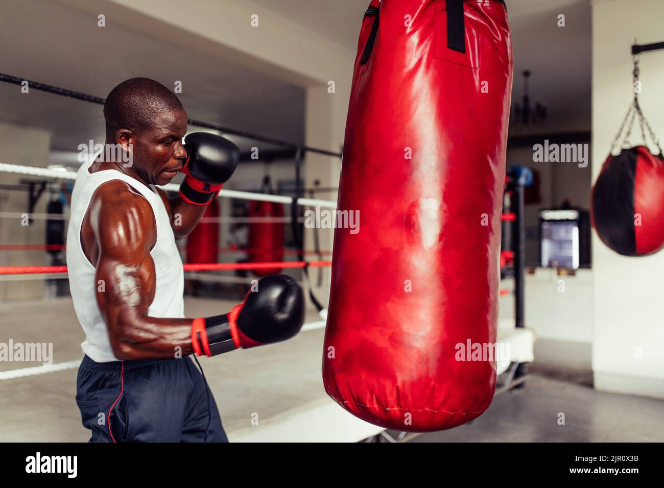 Un jeune boxeur sueur s'entraîner avec un sac de poinçonnage rouge à la salle de gym. Jeune homme sportif pratiquant ses techniques de poinçonnage dans un gymnase de boxe. Banque D'Images