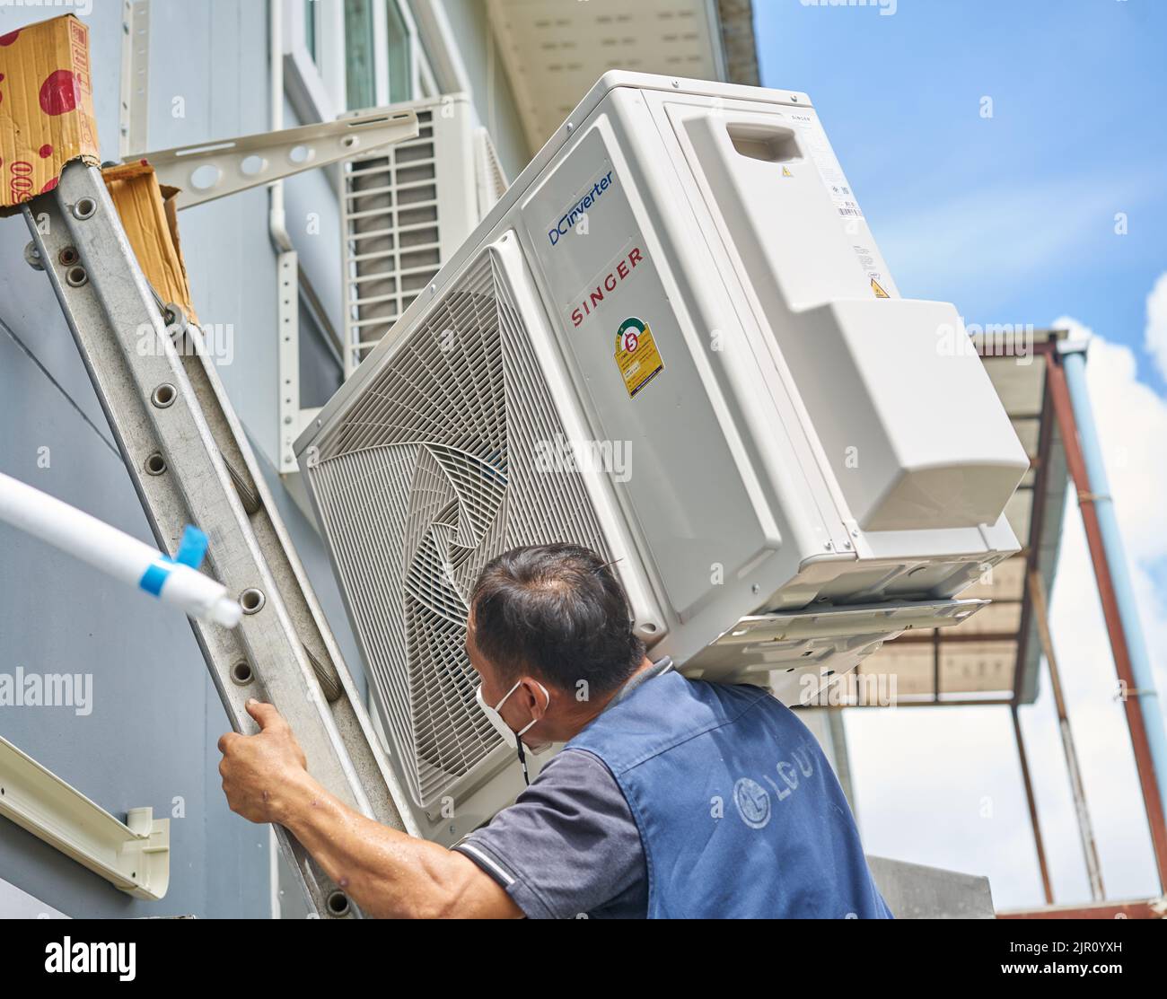 Un homme porte un compresseur de climatisation lourd sur son épaule tout en montant une échelle. Banque D'Images