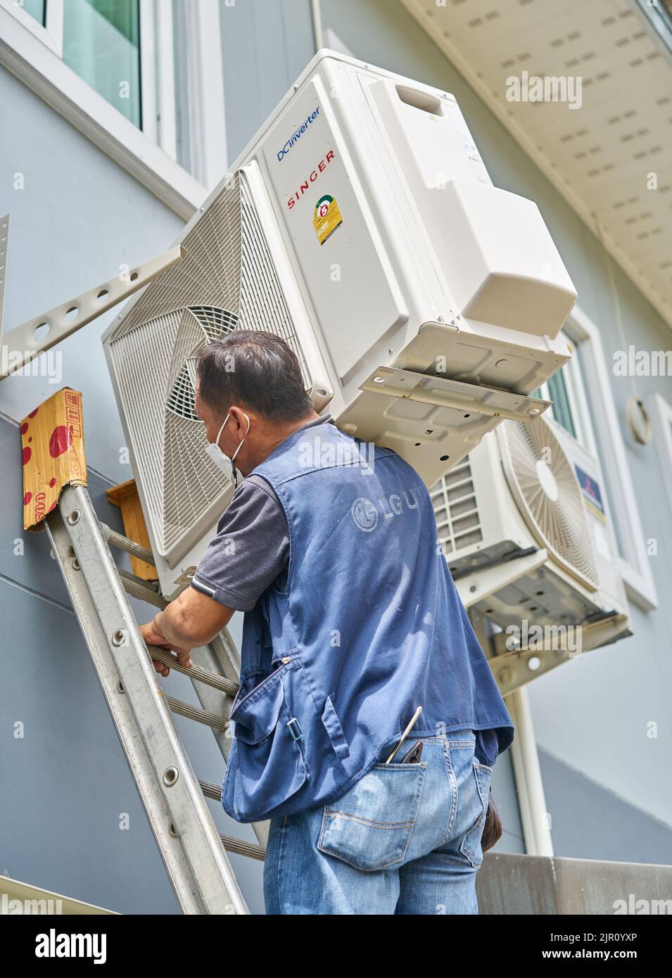 Un homme porte un compresseur de climatisation lourd sur son épaule tout en montant une échelle. Banque D'Images
