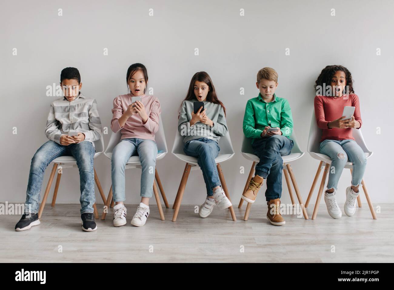 Groupe d'enfants scolaires divers et étonnés utilisant des téléphones intelligents assis à l'intérieur Banque D'Images