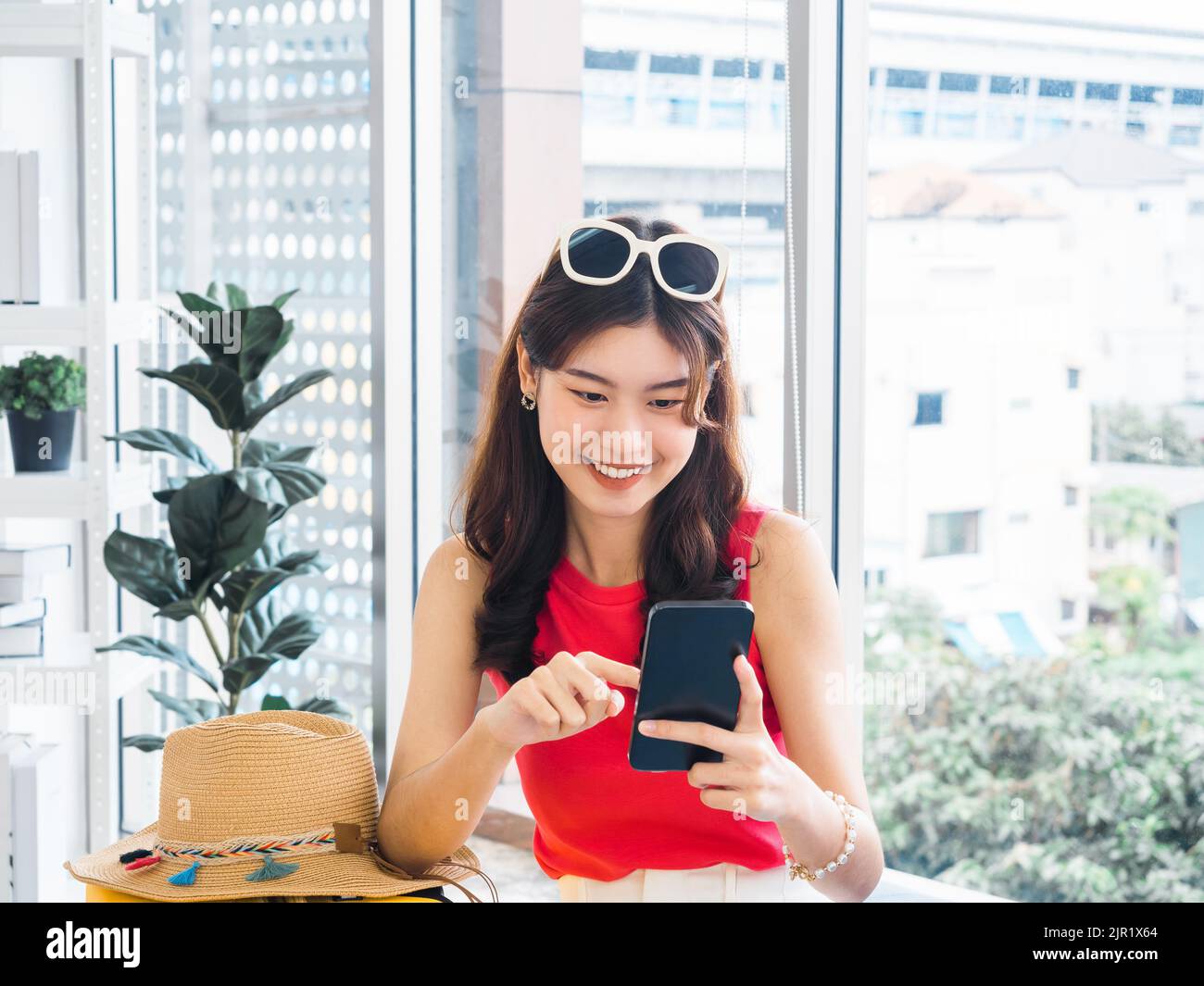 Jeune femme asiatique souriante tenant et utilisant un téléphone mobile intelligent, regardant l'écran. Happy Asian femme vêtu d'une robe sans manches rose, lunettes de soleil, s Banque D'Images