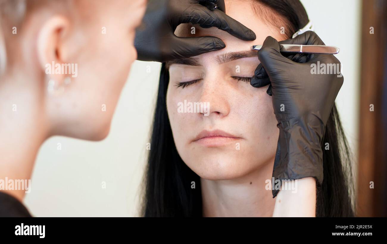 salon de beauté. gros plan, les mains du esthéticien en gants de caoutchouc noir tiennent des pinces et tirent les sourcils. Master corrige la forme des sourcils. Photo de haute qualité Banque D'Images