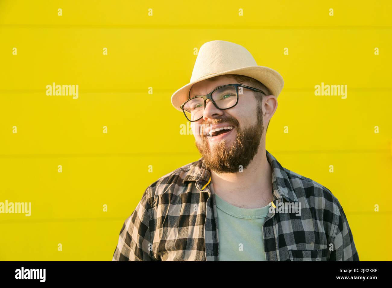 Portrait de rire jeune homme hipster souriant sur le mur jaune magasin arrière-plan extérieur - joli gars à barbe tendance avec chapeau et lunettes debout Banque D'Images