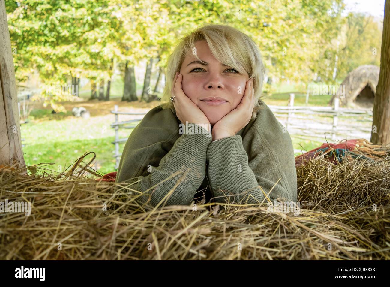 Fille blonde avec des yeux verts dans la nature. Bonne femme d'âge moyen dans une ferme sur une botte de foin lors d'une journée ensoleillée d'automne ou d'été. Banque D'Images
