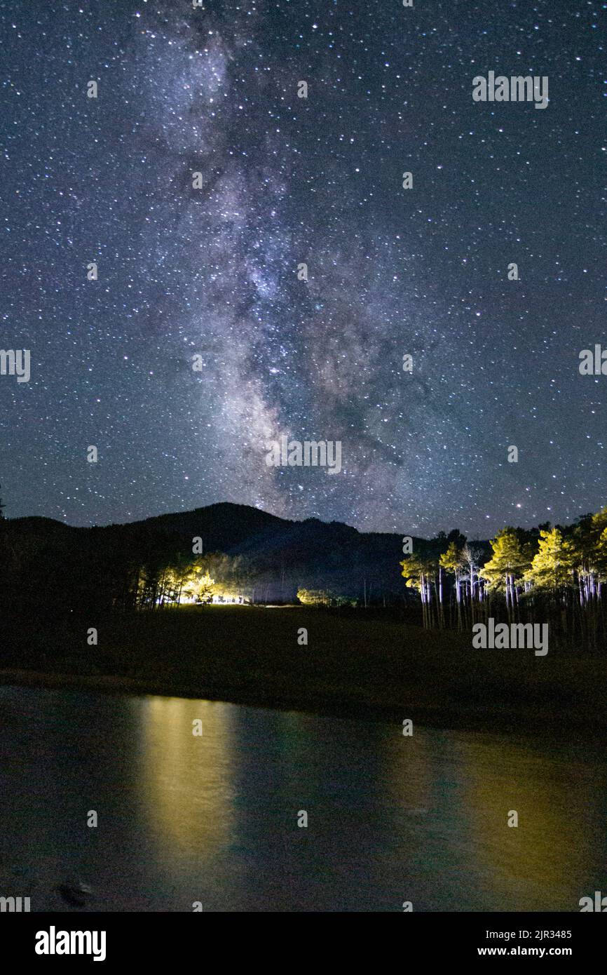 La voie lactée se trouve presque à la verticale au-dessus du lac Isabel dans le sud du Colorado, tandis que les lumières le long de la rive éclairent des peuplements d'épineux Banque D'Images