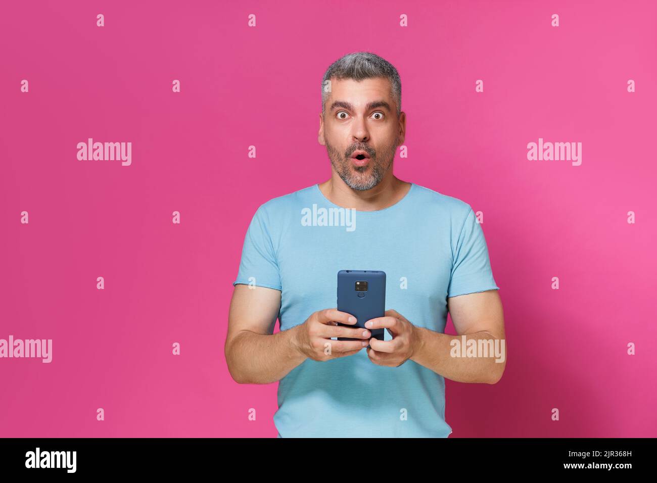 Excitée beau jeune homme adulte, gars 30s 40s en tenue décontractée tenir smartphone lecture de texte ou de navigation en ligne isolé sur fond rose. Mucho homme en t-shirt bleu avec téléphone studio tourné. Banque D'Images