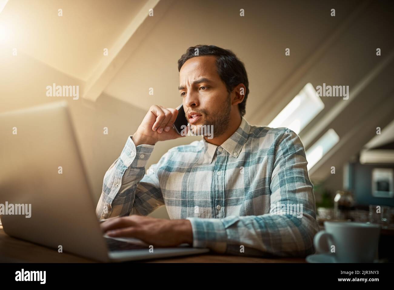 Prendre soin de son café préféré. Un jeune homme parlant sur son téléphone tout en utilisant son ordinateur portable. Banque D'Images