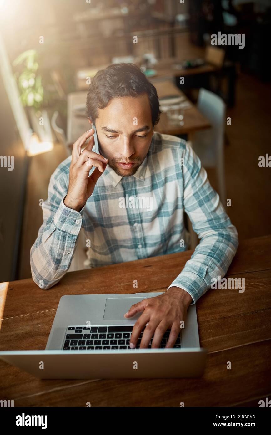 Ne soyez pas occupé, soyez productif. Un jeune homme parlant sur son téléphone tout en utilisant son ordinateur portable. Banque D'Images