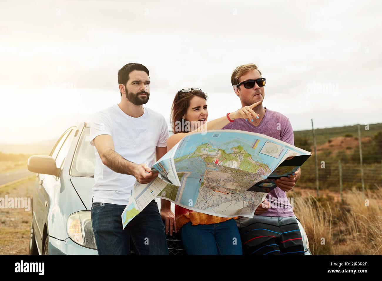 Temps de voyage. Un groupe de jeunes amis tenant une carte routière et planifiant où aller en se tenant à côté d'une route. Banque D'Images