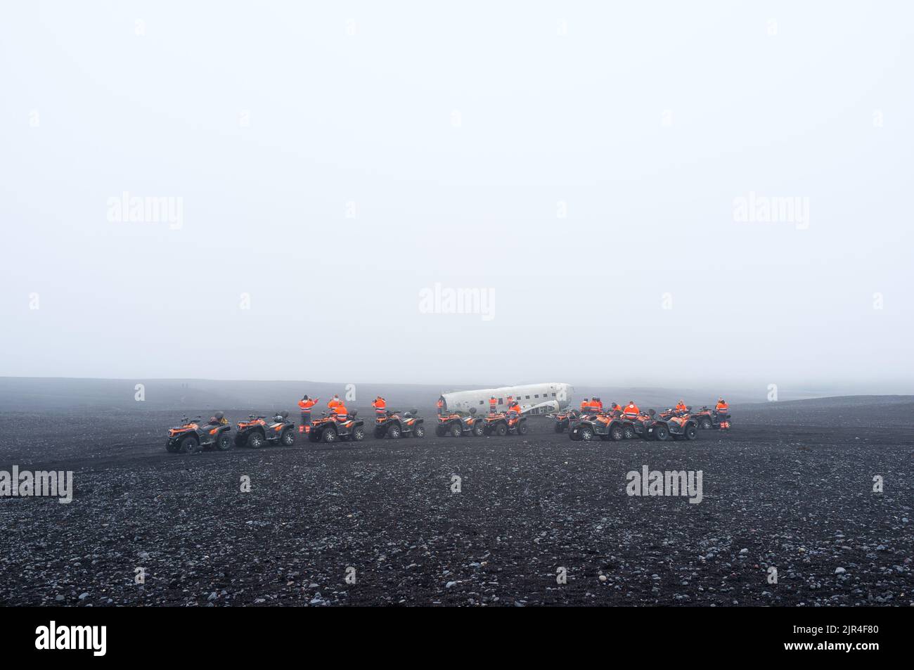 Touristes dans des combinaisons d'orange sur des quads près de l'épave abandonnée d'un avion DC sur Solheimasandur en Islande, un jour brumeux Banque D'Images Touristes dans des combinaisons d'orange sur des quads près de l'épave abandonnée d'un avion DC sur Solheimasandur en Islande, un jour brumeux Banque D'Images